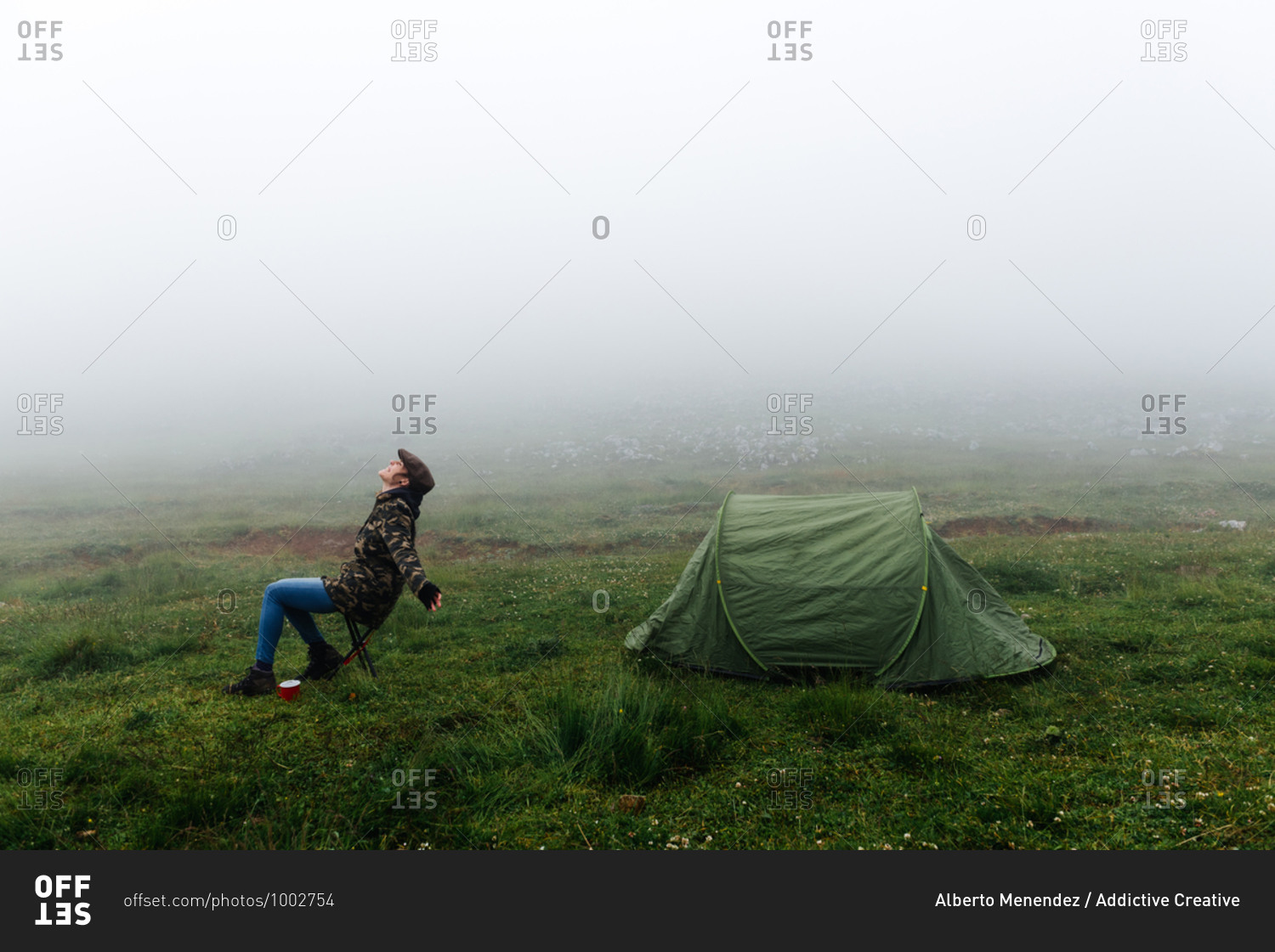 Full length content female traveler in jeans and warm khaki jacket sitting on folding chair near camping tent with cup of hot drink during camping in highlands with closed eyes