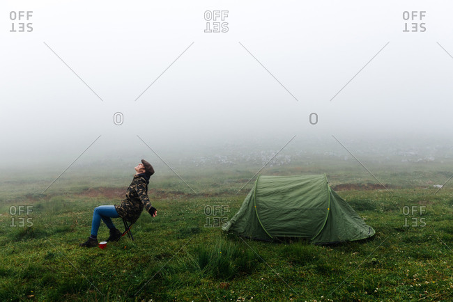 Full length content female traveler in jeans and warm khaki jacket sitting on folding chair near camping tent with cup of hot drink during camping in highlands with closed eyes