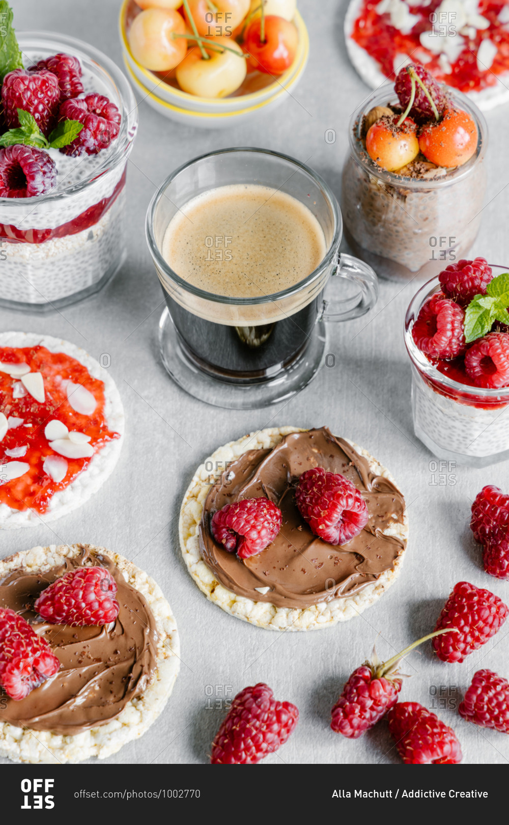 High angle of glass cup of coffee with chia pudding garnished with raspberries and white cherries with wheat crispbreads with nut spread and jam with almond pieces