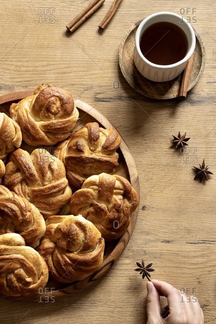 Top view of crop female at wooden table with pile of sweet buns and cinnamon sticks arranged with cup of coffee and star anise