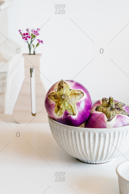 Ceramic bowl with ripe eggplants placed on white table with flowers in modern kitchen