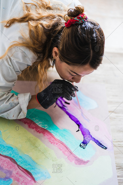 From above of female painter in dirty robe and gloves blowing through thin tube for batik while sitting on wooden floor near colorful abstract painting in art studio