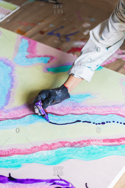From above of crop anonymous female illustrator in dirty gloves creating multicolored abstract painting on large paper sheet holding plastic bottle with paint sitting on wooden floor in workshop