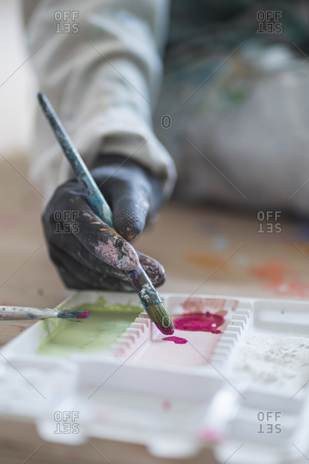 Crop anonymous female illustrator in dirty robe and gloves sitting on floor with paintbrush in front of palette with gouache paints in workshop