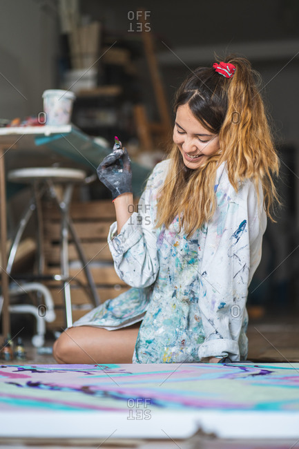 Cheerful barefoot female artist in dirty robe and gloves lying on wooden floor with closed eyes in front of abstract painting in art studio