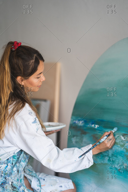 Side view female master in dress smeared with paint painting seascape with paintbrush on round canvas fixed on wooden easel in art studio