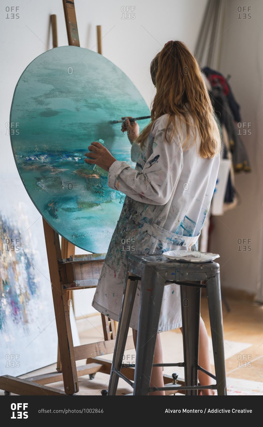 Side view of unrecognizable focused female master in dress smeared with paint painting seascape with paintbrush on round canvas fixed on wooden easel in art studio