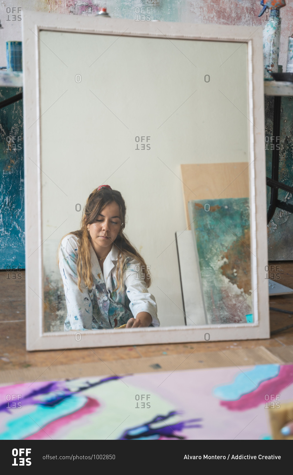 young female artist reflected in mirror painting creative picture with paintbrush and colorful paints on canvas board placed on floor in art studio