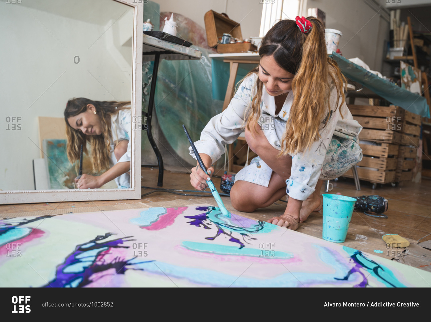 Cheerful barefoot female artist in dirty robe and gloves lying on wooden floor with closed eyes in front of abstract painting in art studio
