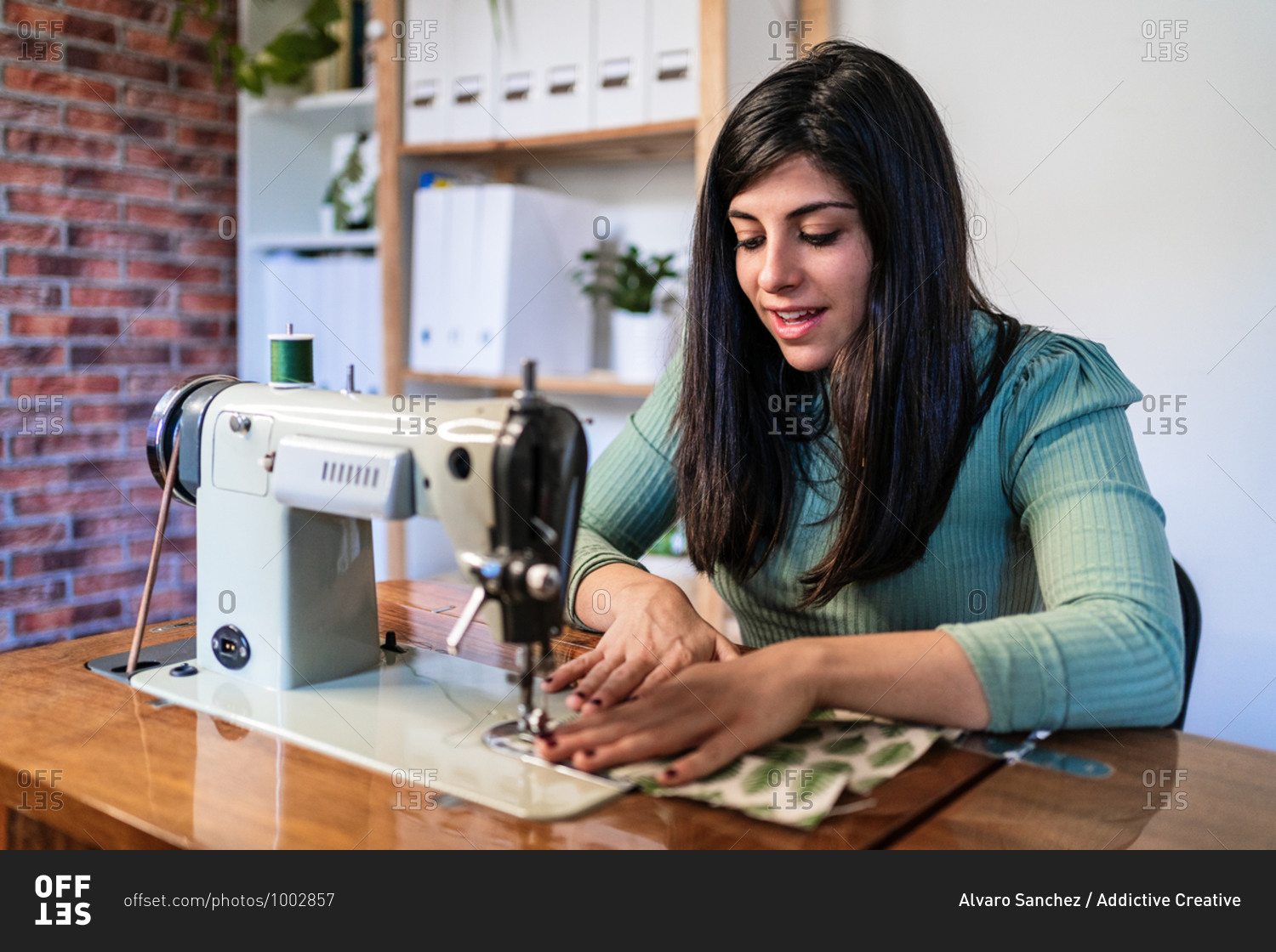 Craftswoman using modern sewing machine while creating soft fabric samples with creative green pattern near lamp in loft style workshop