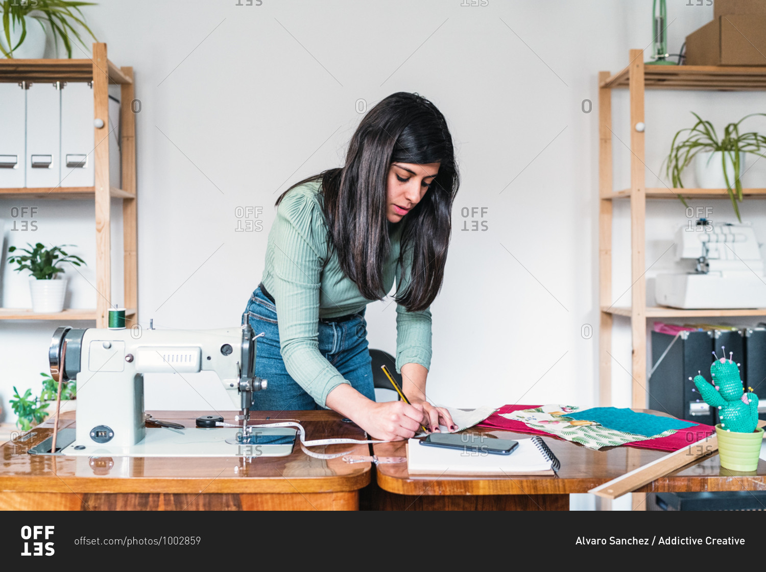 Ethnic female tailor in casual wear taking note on sheet of paper while leaning forward near sewing machine and blank notepad with fabric samples in workshop