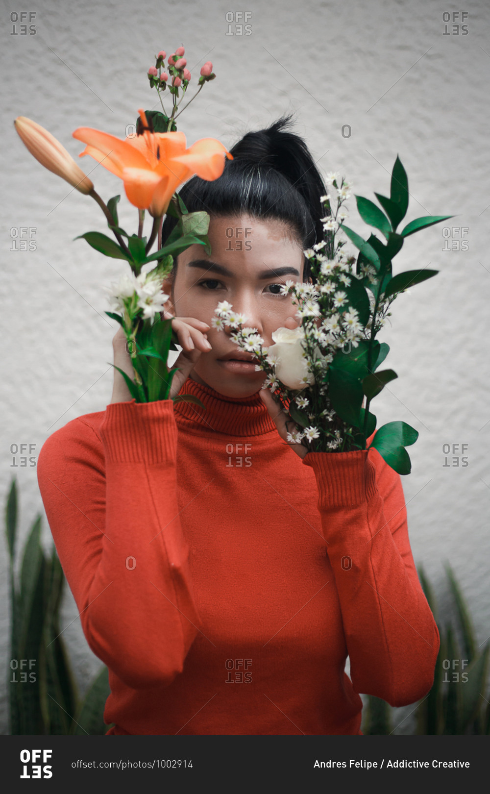 Young unemotional ethnic female with vitiligo in red turtleneck looking at camera while standing with different blooming flowers put in sleeves in apartment