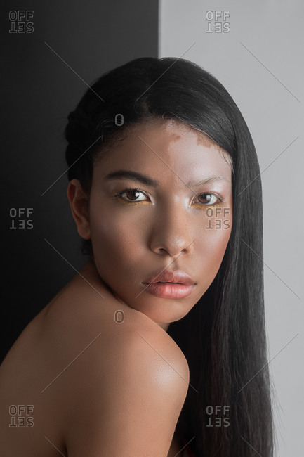 Side view of young sensual emotionless ethnic female with dark hair and vitiligo skin condition looking at camera while standing near black and white wall in flat