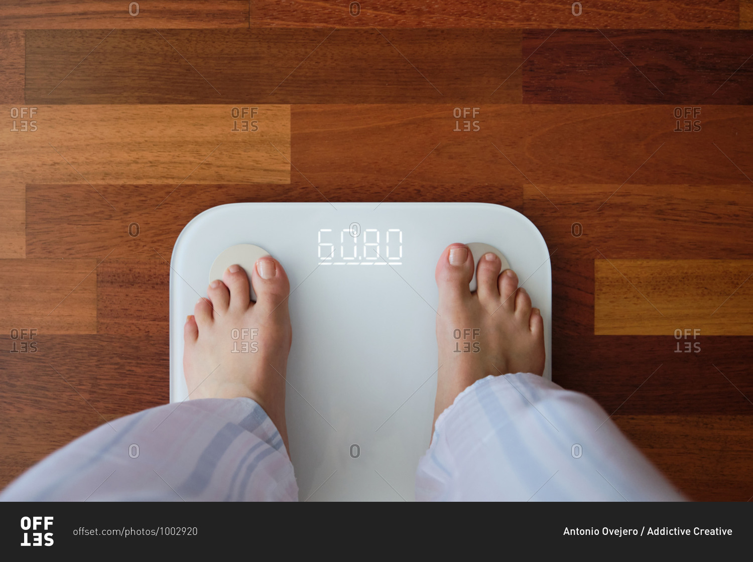Top view crop faceless barefoot female in cozy pajama standing on digital weight and body fat scales with display showing healthy weight of 60 kg on bathroom floor in morning