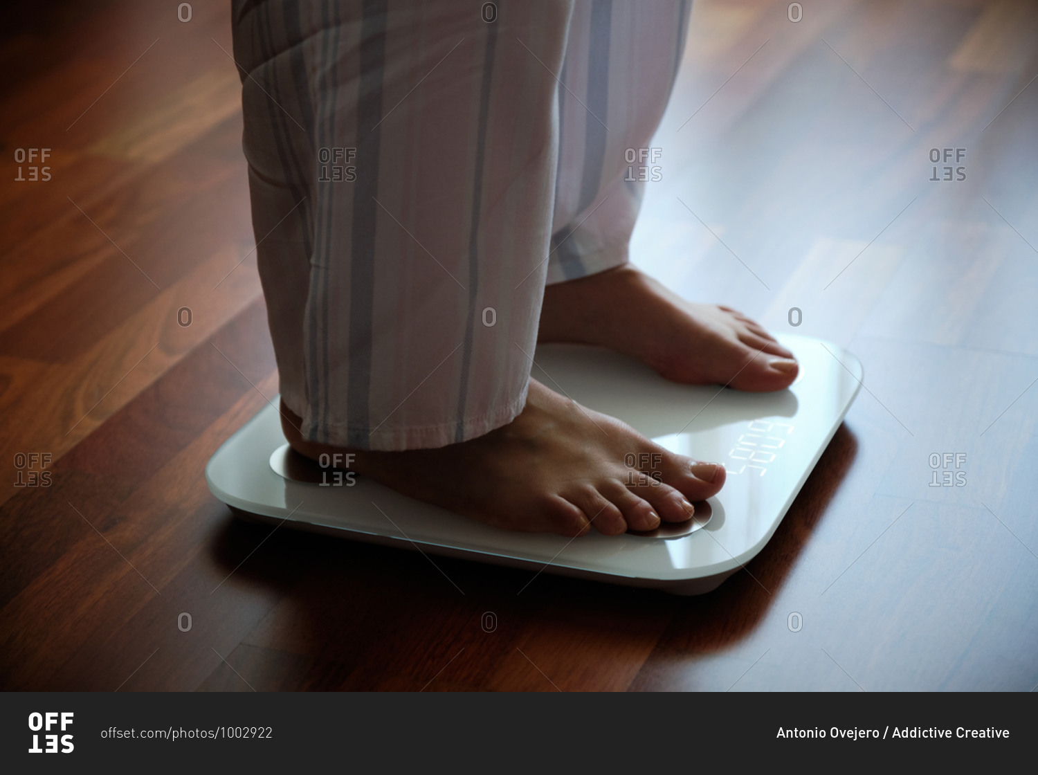 Crop faceless barefoot female in cozy pajama standing on digital weight and body fat scales with display on bathroom floor in the morning