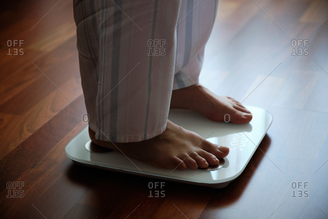 Crop faceless barefoot female in cozy pajama standing on digital weight and body fat scales with display on bathroom floor in the morning
