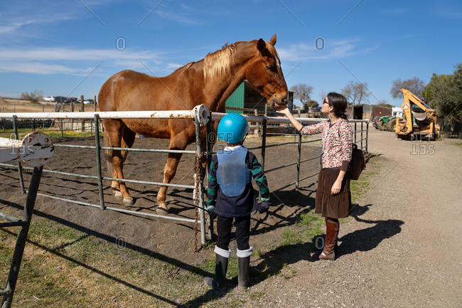 Cheerful woman in casual wear and preteen equestrian in protective helmet standing on sandy arena near fence and caressing chestnut horse