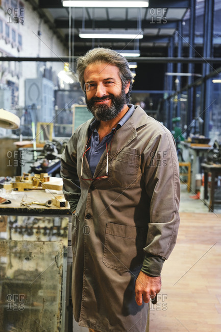 Happy middle aged bearded craftsman in robe with hanging eyeglasses standing and looking at camera in studio
