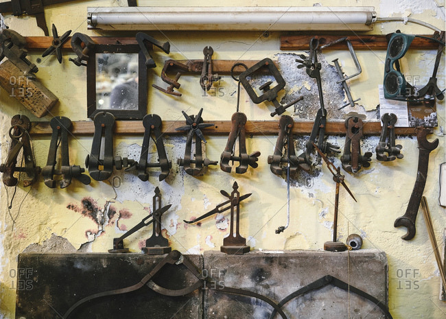Set of various metal jewelry instruments on wooden beam representing pliers and bench pins near saw frames and mirror on concrete weathered wall in studio