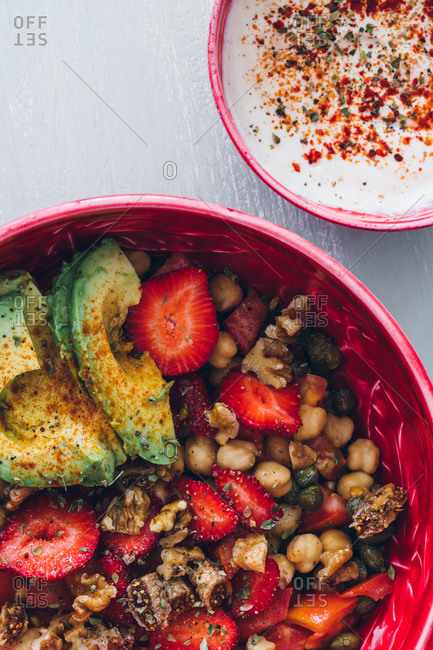Top view composition of crop anonymous female with bowl of yummy healthy chickpeas and avocado salad topped with ripe strawberry and walnuts near bowl with cream sauce and wooden cutleries