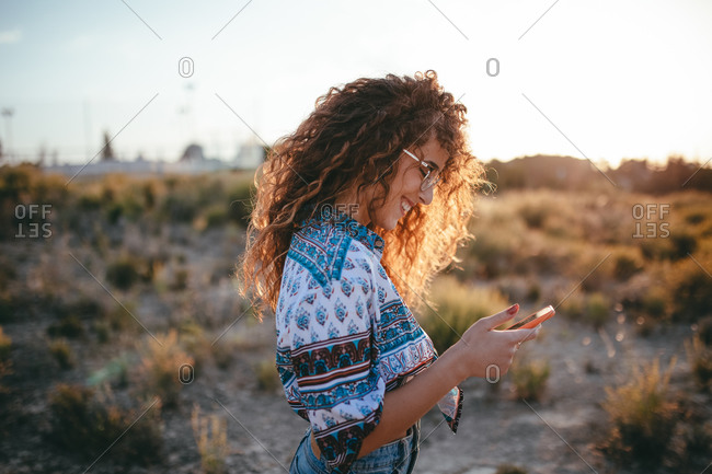 Side view of cheerful female in casual wear and eyeglasses chatting on cellphone while standing near faded grass on uneven terrain in countryside in back lit on sunny day