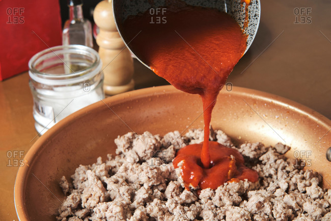Anonymous person pouring homemade tomato sauce into frying pan with fried minced meat while cooking Bolognese sauce in kitchen