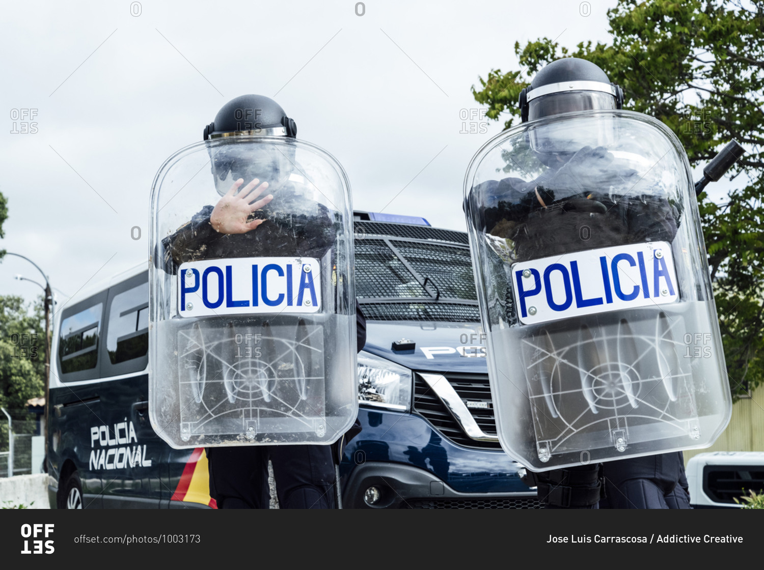 Low angle of anonymous police soldiers in protective uniforms and helmets standing against squad van and defending by riot shields