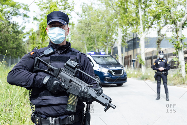 Concentrated police officer in uniform and medical mask looking away while patrolling street with automatic rifle