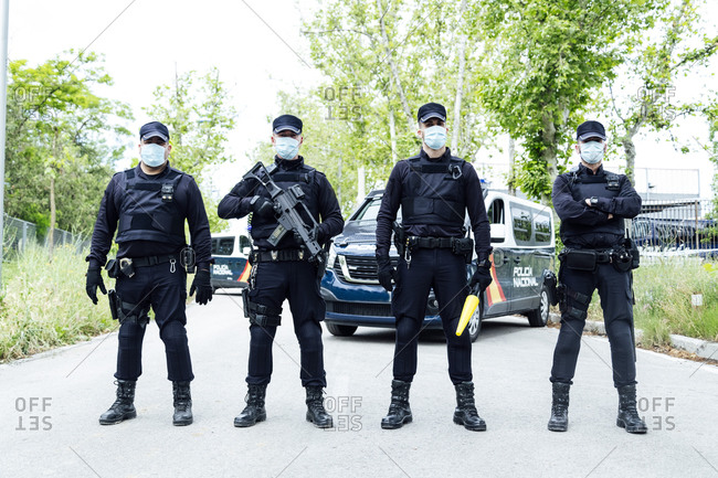 Full body squad of Spanish police officers in protective gears with guns wearing medical masks during patrolling street