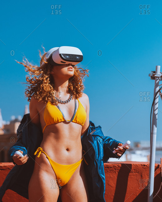 Sensual unrecognizable female traveler in swimwear and VR googles entertaining while watching video with open mouth on roof under blue sky in sunlight