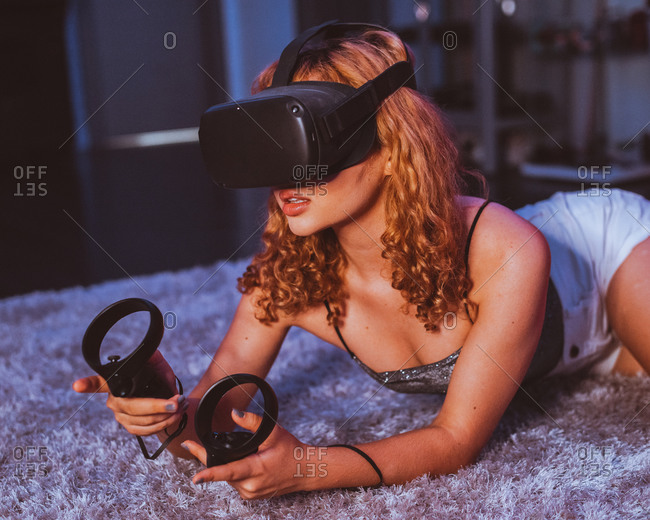 Seductive barefoot anonymous female in casual wear lying with open mouth on soft plaid on bed while playing game with controllers and in goggles of virtual reality