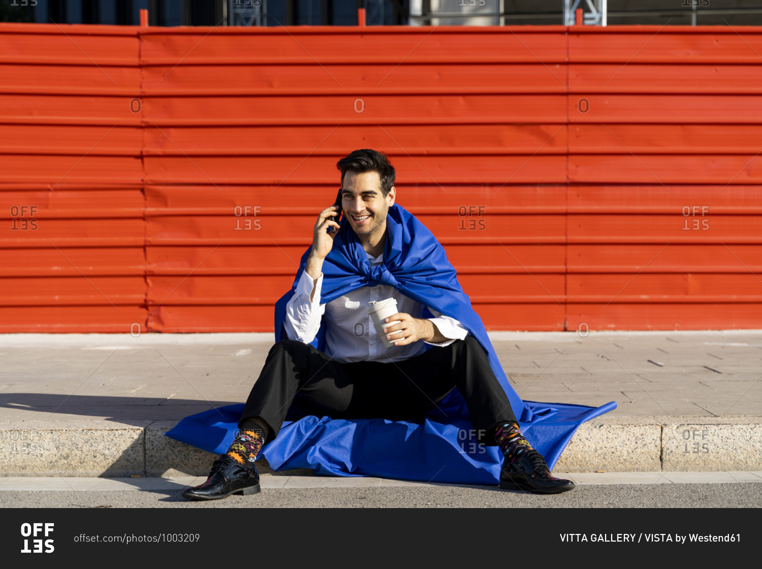 Smiling businessman on the phone wearing superhero cape sitting on curb