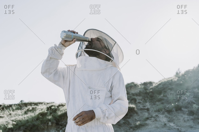 Man wearing a beekeeper dress drinking from thermos flask