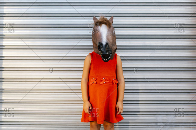 Little girl with a horse\'s head and a red dress in front of  metal shutter