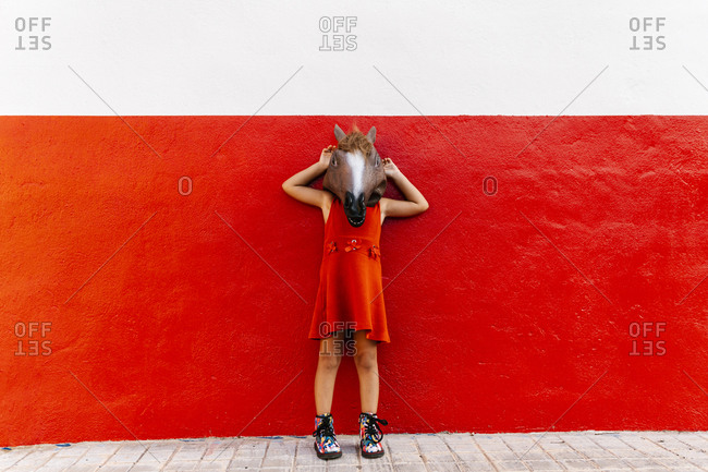 Little girl with a horse\'s head and a red dress leaning on a red and white wall