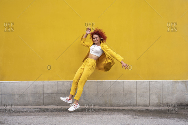 Energetic young woman in yellow suit running and jumping in front of yellow wall