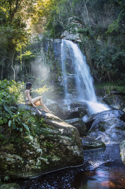 A beautiful young woman sitting in a shaft of sunlight in front of a pristine waterfall in the rainforest, Brazil, South America