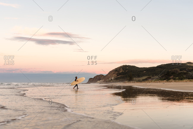 Boy carrying a surfboard at sundown on the coast of New Zealand