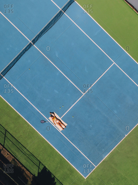 Aerial view of a blond woman sunbathing on a tennis court with a tennis racket