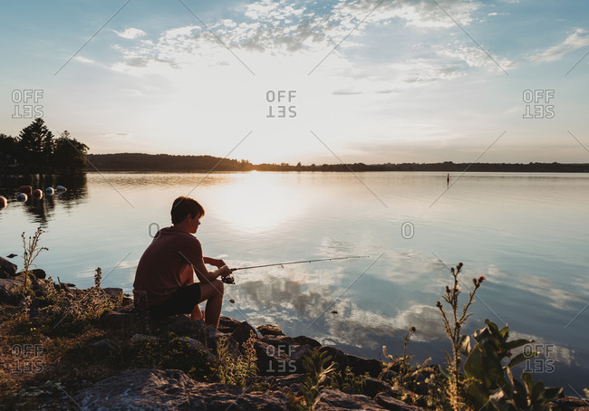 Adolescent boy fishing on shore of lake at sunset in Ontario, Canada.