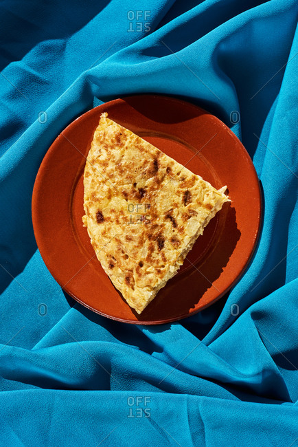 High angle view of a portion of typical Spanish omelet in a brown earthenware plate, on a wrinkled blue fabric