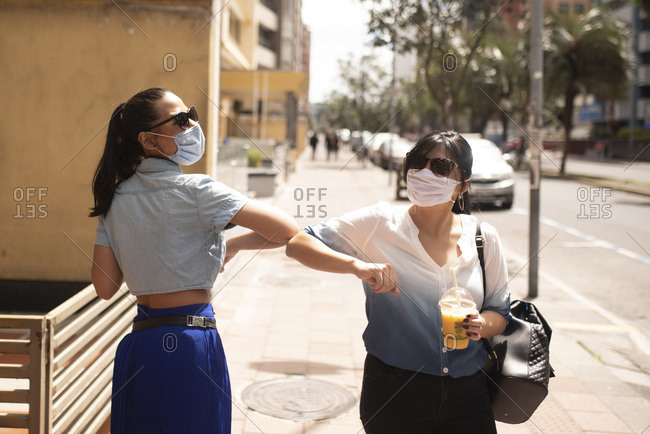 Female friends wearing sunglasses and masks giving elbow dump while standing in city