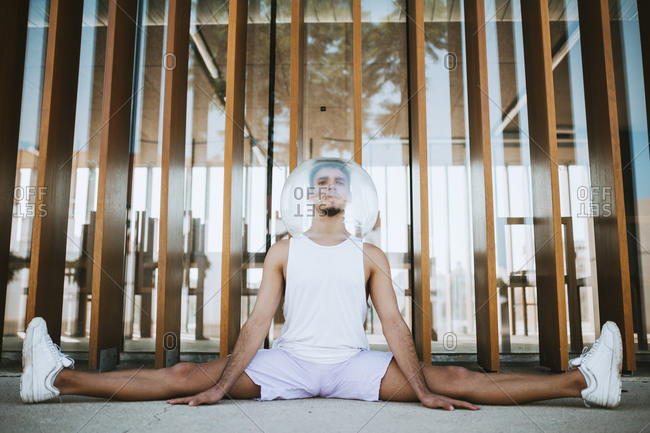 Young man wearing glass container on head with legs apart exercising against built structure