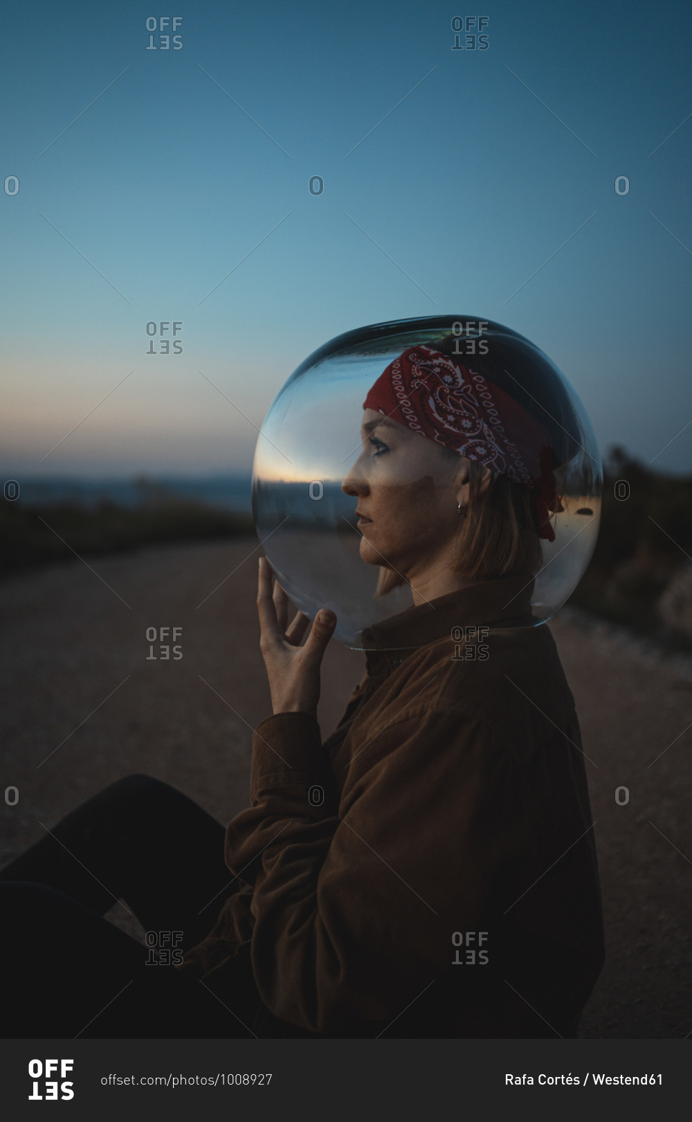 Woman with a fish bowl on her head sitting on a road in the countryside
