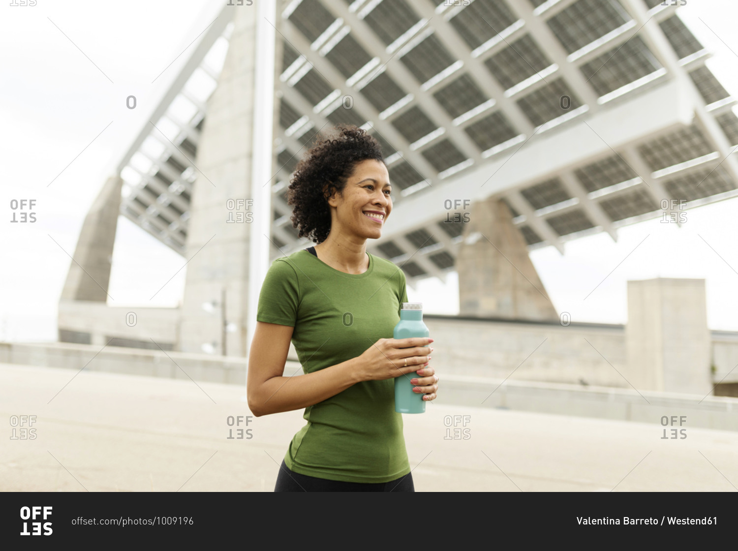 Smiling woman holding water bottle looking away while standing against built structure in city