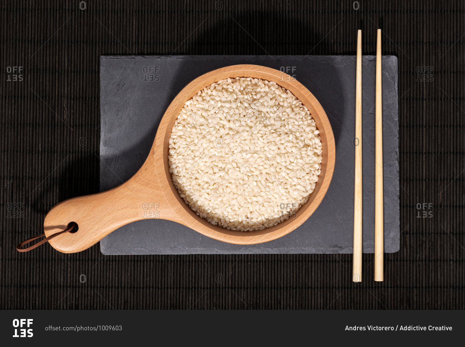 Top view of wooden bowl with raw rice and bamboo chopsticks placed on black slate board in restaurant