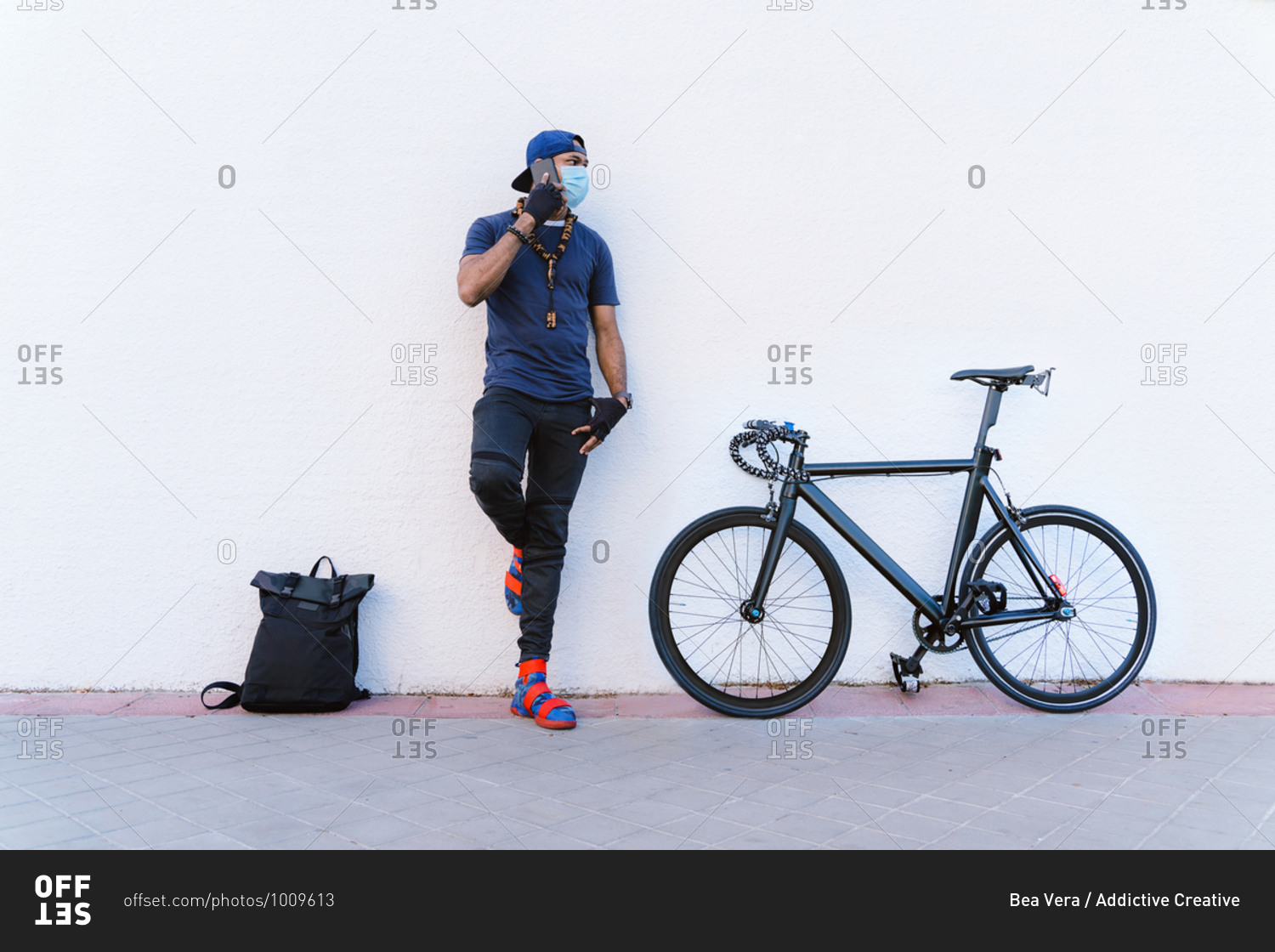 Full body young African American man in medical mask speaking on mobile phone while standing near backpack and bicycle near white stone wall on urban street