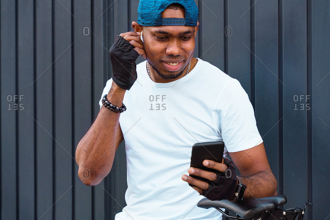 Positive young African American male biker with bracelets and cap adjusting earbuds and choosing playlist on smartphone while standing with bicycle against gray metal wall