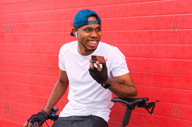 Positive young African American male biker with bracelets, cap and using earbuds while speaking on smartphone standing with bicycle resting against red wall on street