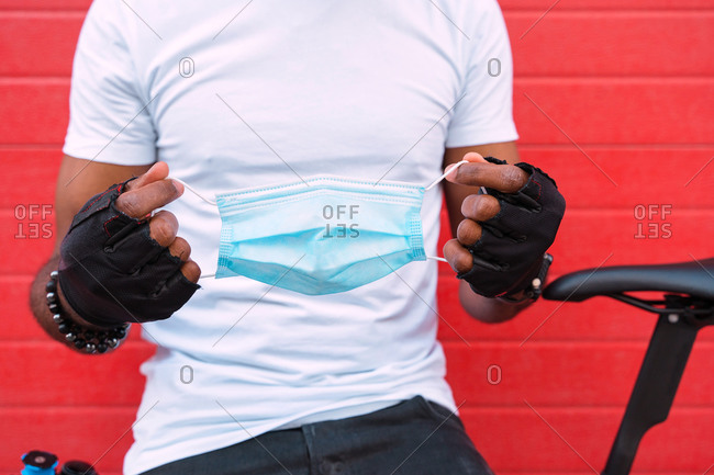 Crop anonymous black male in biker gloves and bracelet holding disposable medical mask while standing with bicycle against red wall on street